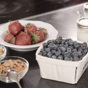 Ingredients for Overnight Groats Breakfast Recipe for Macular Degeneration - A white bowl of fresh strawberries, a basket of fresh blueberries, a glass bowl of almonds, a glass bowl of walnuts, and two mason jars with milk.