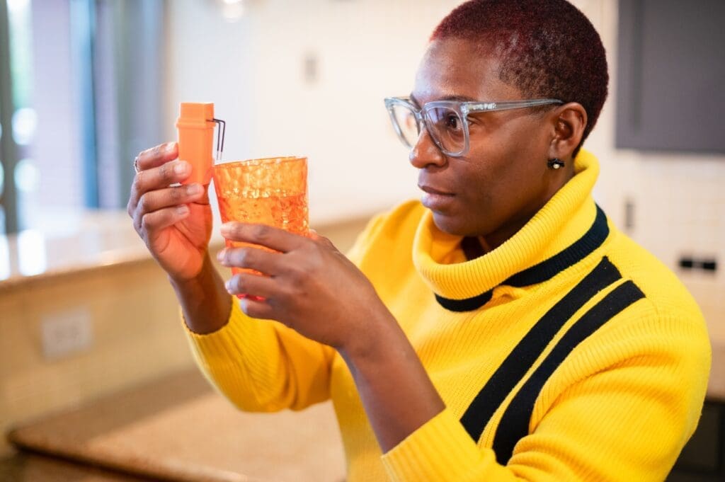 Woman holing a cup and a low vision device to alert when liquid has reached the fill line. She's wearing glasses and a bright yellow shirt with two black stripes.